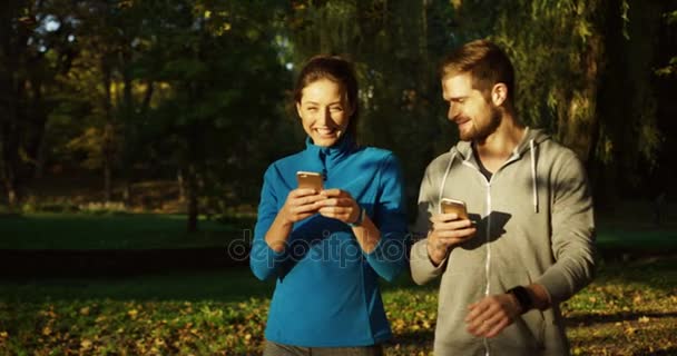 Νεαρό ζευγάρι από joggers περίπατο στο όμορφο πάρκο φθινόπωρο, γελώντας και να δέσει με ταινία τα κινητά τους τηλέφωνα. Ηλιόλουστη μέρα. Εξωτερική — Αρχείο Βίντεο