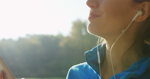 Mujer bonita con auriculares relajándose en el parque mientras escucha la música en el teléfono después de correr. Clima soleado. Afuera. De cerca. Retrato — Vídeos de Stock