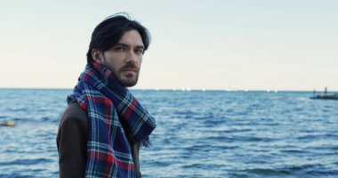 Portre çekim gözlük ve denize bakarak ve sahilde kafasını kameraya dönüm motly eşarp melankolik yakışıklı bir adam. Bulutlu gün. Açık