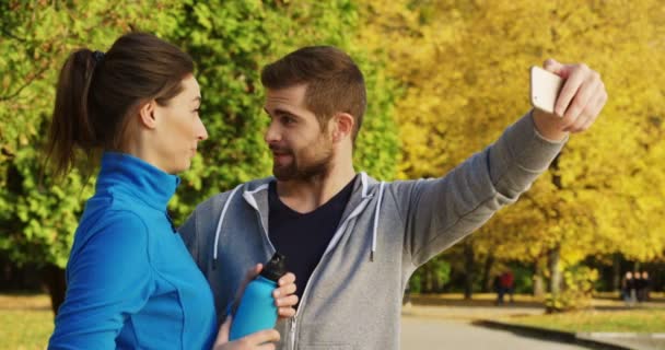 Joven pareja deportiva sonriente teniendo video chat, saludando y enviando besos de golpe en el teléfono inteligente en el soleado parque a principios de otoño. Exterior . — Vídeos de Stock