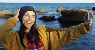 Esmer womanmaking büyüleyici akıllı telefonunu kayalar ve deniz arka plan üzerinde güzel selfies gülümsedi. Soğuk ama güneşli hava. Açık havada