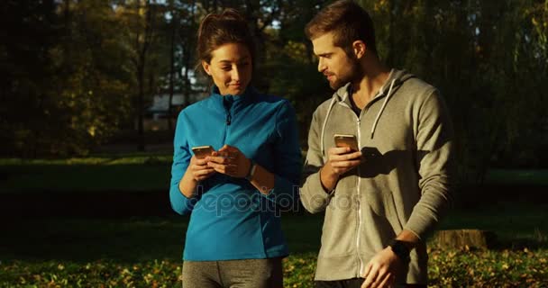 Joven pareja deportiva de corredores paseando por el hermoso parque de otoño, coqueteando y grabando en sus teléfonos inteligentes. Día soleado. Al aire libre — Vídeo de stock
