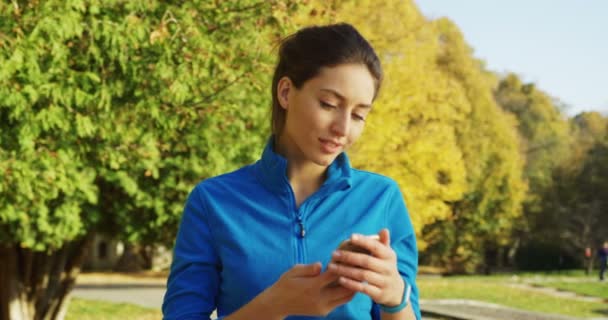 Досить молода жінка біжить в синьому светрі, знімаючи на телефоні і дивлячись на годинник і на стороні в парку, перш ніж бігти. Сонячна гарна погода. На відкритому повітрі. Портрет знятий — стокове відео