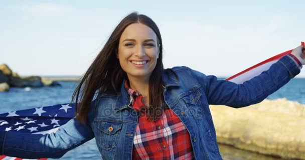 Young charming woman standing at the sea shore and holding a waving American flag.Then wrapping in it and smiling into the camera. Outside — Stock Video