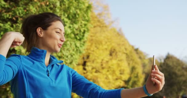 Atractiva mujer sonriente en suéter azul posando y tomando selfies en el parque después de correr. Clima soleado. Afuera. Retrato — Vídeos de Stock