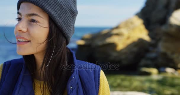 Крупним планом красива молода жінка в капелюсі дивиться збоку, а потім повертає голову до камери на кам'янистому морському фоні. Холодний сонячний і вітряний день. Портрет знятий. На відкритому повітрі — стокове відео