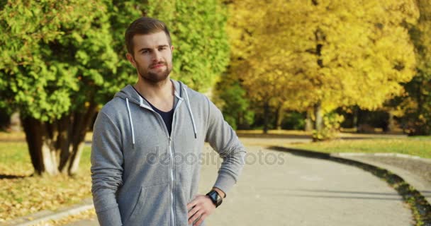 Porträtt av unga kaukasiska härlig sportiga mannen jogger ser kameran och leende i parken i tidigt faller. Utanför. Soligt väder — Stockvideo