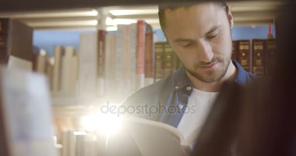 Primer plano del joven guapo leyendo un libro entre estantes con libros en la biblioteca. Un retrato. De interior — Vídeos de Stock