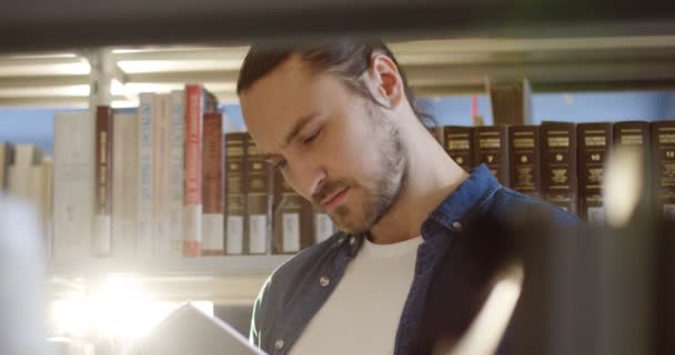 Portret shot van de jonge mannelijke aantrekkelijke student lezen van een boek onder planken met boeken in de bibliotheek. Close-up. Binnenkant — Stockvideo