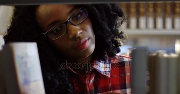 Close up da jovem afro-americana bonita mulher encaracolada olhando através dos livros nas prateleiras da biblioteca. Tiro de retrato. Interior — Vídeo de Stock