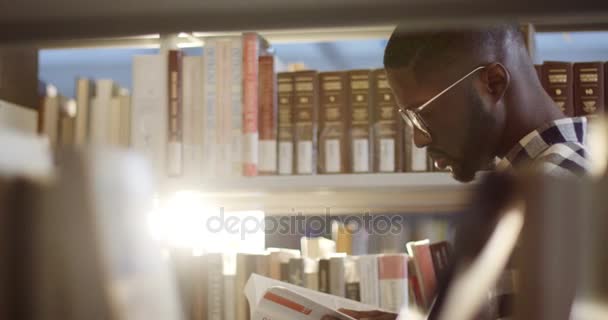 Retrato del lado del joven afroamericano en gafas volteando páginas del libro mientras está de pie entre los estantes de los libros en la biblioteca. De cerca. Al aire libre — Vídeo de stock