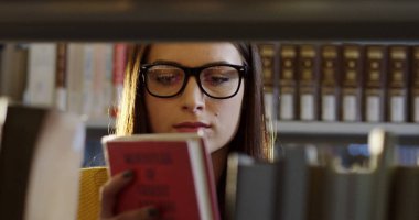 Gözlük chosing genç çekici kadın öğrencinin yakın bir kitap kitaplar kütüphane raflarında arasında. Portre vurdu. Kapalı