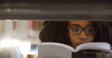 Kütüphanede kitap rafları arasında kitap okuma gözlükleri Afrikalı-Amerikalı genç kıvırcık kadın öğrencinin yakın çekim. Portre. İçinde