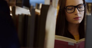 Kütüphanede kitap rafları arasında kitap okuma gözlükleri beyaz genç güzel kadın öğrencinin yakın çekim. Portre. Kapalı