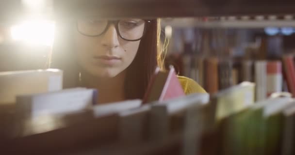 Primer plano de la joven hermosa estudiante en gafas buscando un libro entre los libros en los estantes de la biblioteca. Un retrato. De interior — Vídeos de Stock