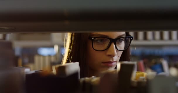 Närbild på den unga ganska kvinnlig studenten i glasögon vänder sidor stående bland böcker på hyllorna i biblioteket. Porträtt. Inomhus — Stockvideo