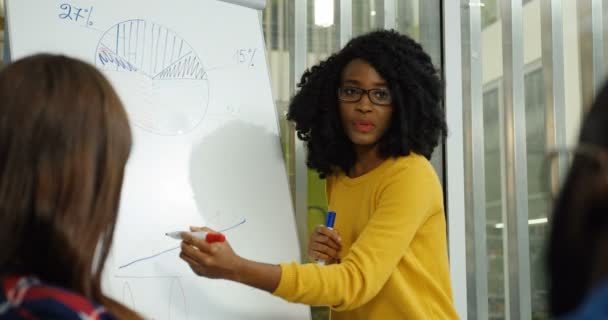 Retrato de la joven afroamericana rizada en gafas teniendo una discusión con sus colegas mientras dibuja en la pizarra gráficos y gráficos en la bonita sala de oficina. De interior — Vídeo de stock