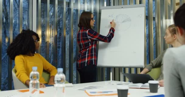 Jóvenes carreras mixtas compañeras de trabajo consultando juntas sobre el nuevo proyecto de puesta en marcha dentro de la bonita sala de oficina y una de ellas dibujando en la pizarra . — Vídeos de Stock