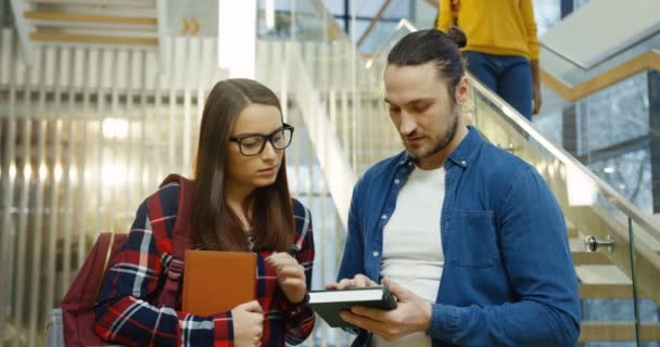 Junges attraktives Studentenpaar, das auf dem Smartphone spricht und zusieht, während der Mann darauf blättert. im Inneren des großen modernen Gebäudes. — Stockvideo