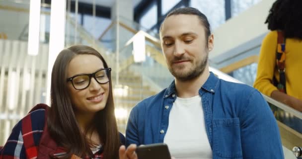 Young attractive woman and young handsome man talking and laughing at something they watching on the smart phone while standing in the nice big modern building. Indoor — Stock Video