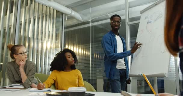 Ein junges gemischtes Team von Büromitarbeitern diskutiert das neue Start-up und ein junger Mann zeichnet Diagramme auf der Tafel im Büro. drinnen — Stockvideo