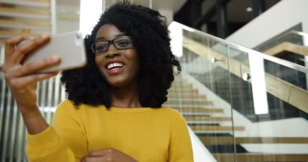 Close up da jovem afro-americana bem-parecido mulher em óculos ter vídeo chat no smartphone branco no agradável salão moderno. Tiro de retrato. Interior — Vídeo de Stock
