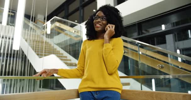 Sonrió hermosa mujer joven afroamericana con el pelo rizado de pie en las escaleras y hablando por el teléfono móvil. Adentro. — Vídeo de stock