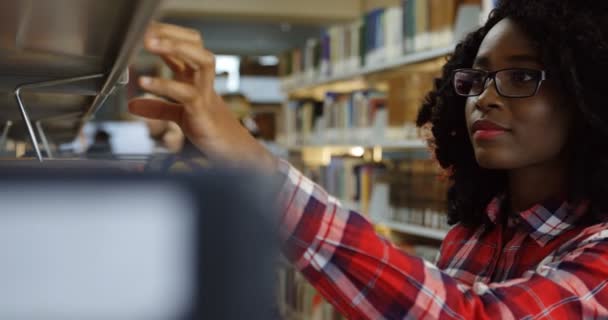 Nahaufnahme der afrikanisch-amerikanischen lockigen Frau mit Brille, die in der Bibliothek steht und sich ein Buch aussucht, als es zu öffnen und zu lesen. Porträt. drinnen — Stockvideo