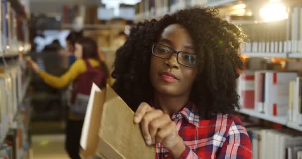 Primer plano de la joven estudiante afroamericana rizada en gafas leyendo un viejo libro entre los estantes de libros en la biblioteca. Retrato. De interior — Vídeos de Stock