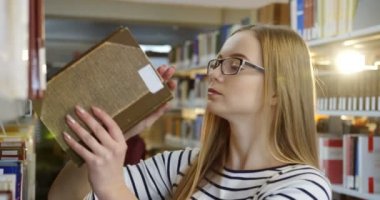 Kütüphanede duran ve açma ve okuma daha bir kitap seçerek sarı saçlı gözlüklü beyaz güzel kadın portre vurdu. Yakın çekim. Kapalı
