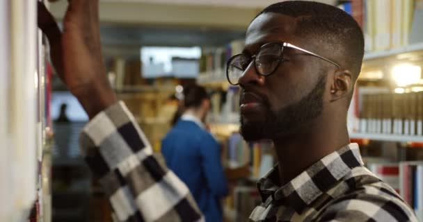 Close-up do afro-americano jovens estudantes em óculos de pé na passagem da biblioteca e à procura de um livro, do que abri-lo. Tiro de retrato. Interior — Vídeo de Stock