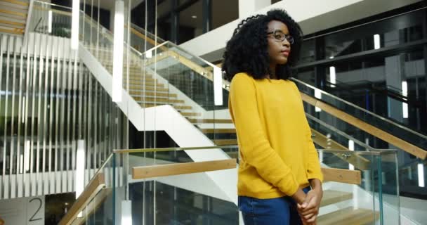 젊은 아프리카계 미국인 여자 곱슬 머리와 안경에는 camra 포즈, 그녀의 손에 건너와 도시 builduing 홀에 서 있는 동안 웃 고. 세로 촬영. 실내 — 비디오