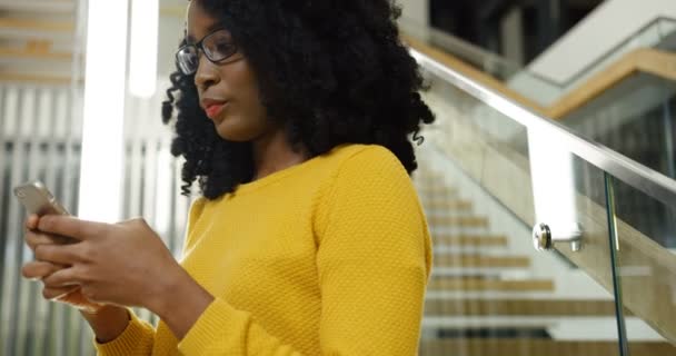 Jovem mulher afro-americana atraente encaracolado em óculos sorrindo e conversando no telefone inteligente perto de grandes escadas modernas. Fecha. Tiro de retrato. Interior — Vídeo de Stock