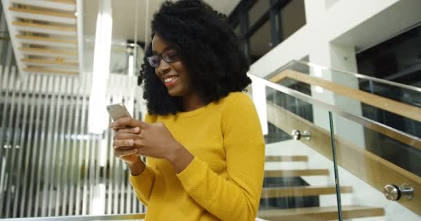 Joven mujer afroamericana rizada en gafas riendo mientras envía mensajes de texto y recibe mensajes en su teléfono inteligente cerca de grandes escaleras urbanas en el pasillo. Un retrato. En interiores — Vídeo de stock