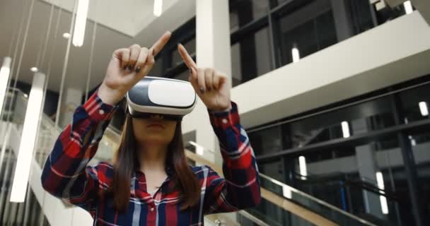 Portrait de la jeune femme en lunettes VR et chemise à carreaux défilant, filmant et zoomant dans les airs pendant un casque VR. Grand hall moderne sur le fond. Ferme là. Intérieur — Video