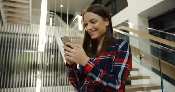 Młoda kobieta uroczy w plaid shirt, śmiejąc się, podczas gdy tekstylny i dostawanie wiadomości w swoim smartfonie w pobliżu dużych miejskich schody w hali. Portret strzał. Wewnątrz — Wideo stockowe