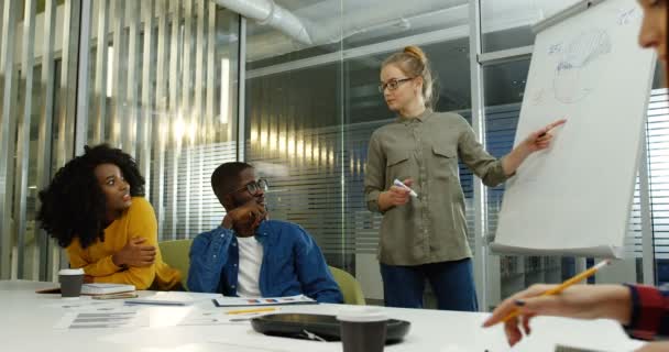 Mladá běloška v brýlích vysvětlující plán svého projektu práce tím, že kreslí na palubě a její mladé kolegy, poslouchat ji. Smíšené rasy tým studentů. Vnitřní — Stock video