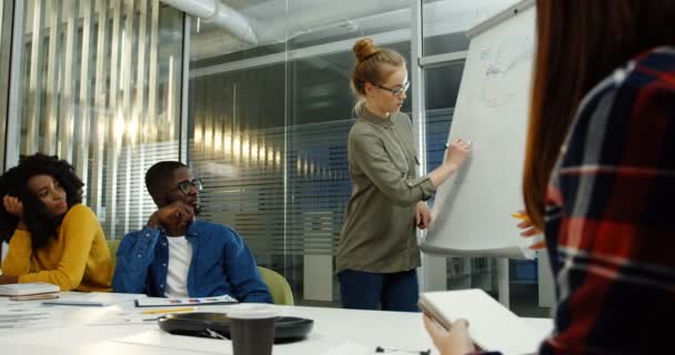 Команда молодых работников смешанных рас обсуждает новый стартап-план и одну молодую женщину, рисующую графики на доске в современной офисной комнате. Внутри — стоковое видео