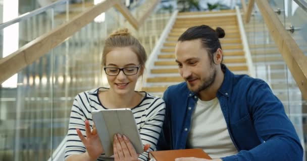 Jovem branco casal atraente de estudantes sentados nas escadas grandes e mulher loira mostrando algo no dispositivo tablet para seu amigo masculino. Sala moderna. Interior — Vídeo de Stock