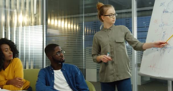 Joven estudiante rubia en gafas explicando un proyecto de estudio en la pizarra y sus amigos compañeros de grupo escuchándola. Grupo multiétnico de estudiantes. En interiores — Vídeos de Stock