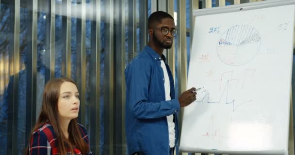 Команда смешанных рас юных студентов проводит мозговой штурм, обсуждая свой план проекта на доске с графикой в удобном классе. В помещении . — стоковое видео