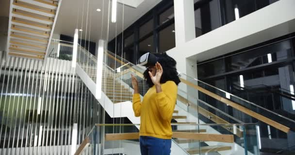 Mujer afroamericana con el pelo rizado de pie en el medio del bonito edificio moderno y con auriculares VR, riendo y grabando en el aire. En interiores — Vídeo de stock