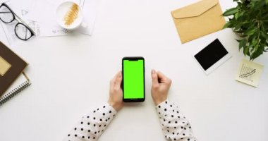 Üstten Görünüm beyaz Ofis Masası ve yeşil ekran ve üzerinde taping eller ile siyah smartphone. Dikey. Yanında başı işleri. Chroma anahtar.