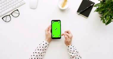 Üstten Görünüm beyaz Ofis Masası ve yeşil ekran ve üzerinde taping eller ile siyah smartphone. Dikey. Yanında başı işleri. Chroma anahtar. Ttracking hareket