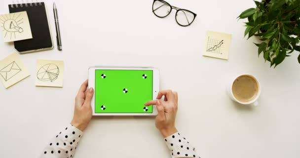 Vue de dessus sur le bureau et la tablette blanche avec écran vert et mains féminines collant dessus. Horizontal. Des affaires de bureau et du café à côté. Clé chromatique. Mouvement de suivi 