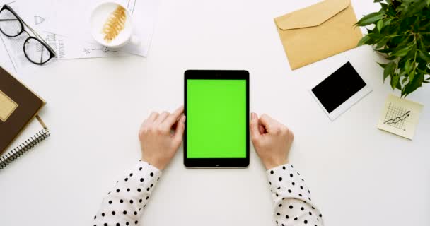 Ovanifrån på vita skrivbord och svart surfplatta med grön skärm och kvinnliga händer tejpa på den. Lodrät. Kontor grejer bredvid. Färgtransparens. — Stockvideo