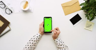 Üstten Görünüm beyaz Ofis Masası ve yeşil ekran ve üzerinde taping eller ile siyah smartphone. Dikey. Yanında başı işleri. Chroma anahtar. Ttracking hareket