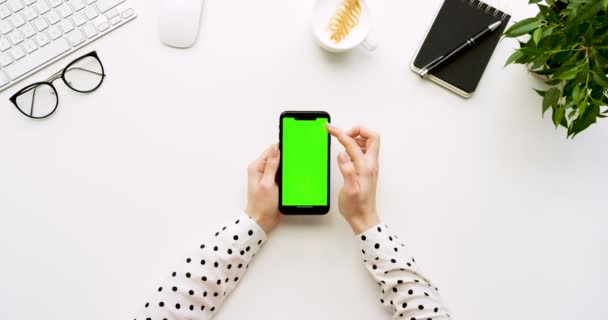 Вид зверху на білий офісний стіл і чорний смартфон з зеленим екраном і жіночими руками, прикріпленими на ньому. Вертикально. Офісні речі поруч. Клавіша Chroma . — стокове відео