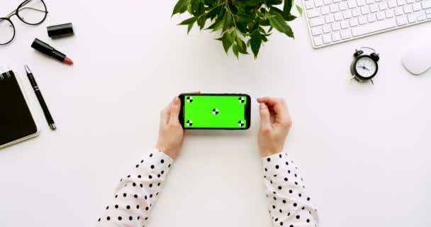 Ovanifrån på vita skrivbord och svart smartphone med grön skärm och kvinnliga händer tejpa på den. Horisontella. Kontor grejer bredvid. Färgtransparens. Spåra rörelse — Stockvideo