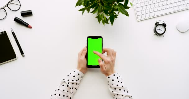 Ovanifrån på vita skrivbord och svart smartphone med grön skärm och kvinnliga händer tejpa på den. Lodrät. Kontor grejer bredvid. Färgtransparens. — Stockvideo